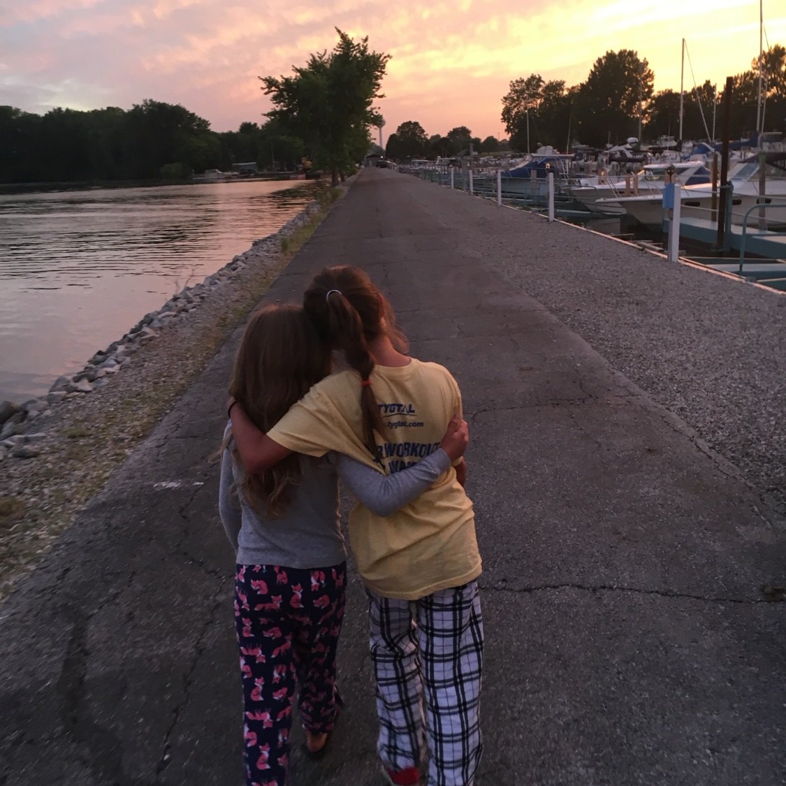 Two granddaughters in pajamas walking arm in arm down the marina pier at sunset, boats tied up alongside